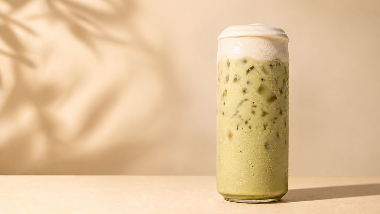 Iced pistachio latte in a tall glass on a warm sand-coloured table, a palm shadow falling across the wall behind