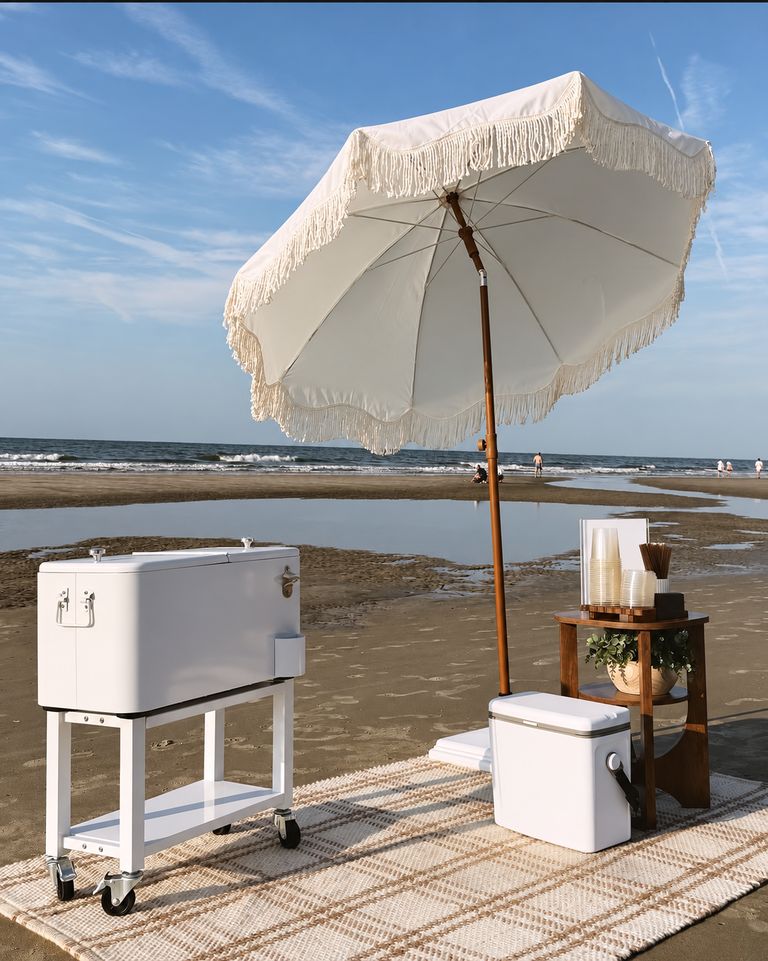 Beach pop-up with a fringed umbrella, a rolling cooler cart and a wooden service table by the shoreline
